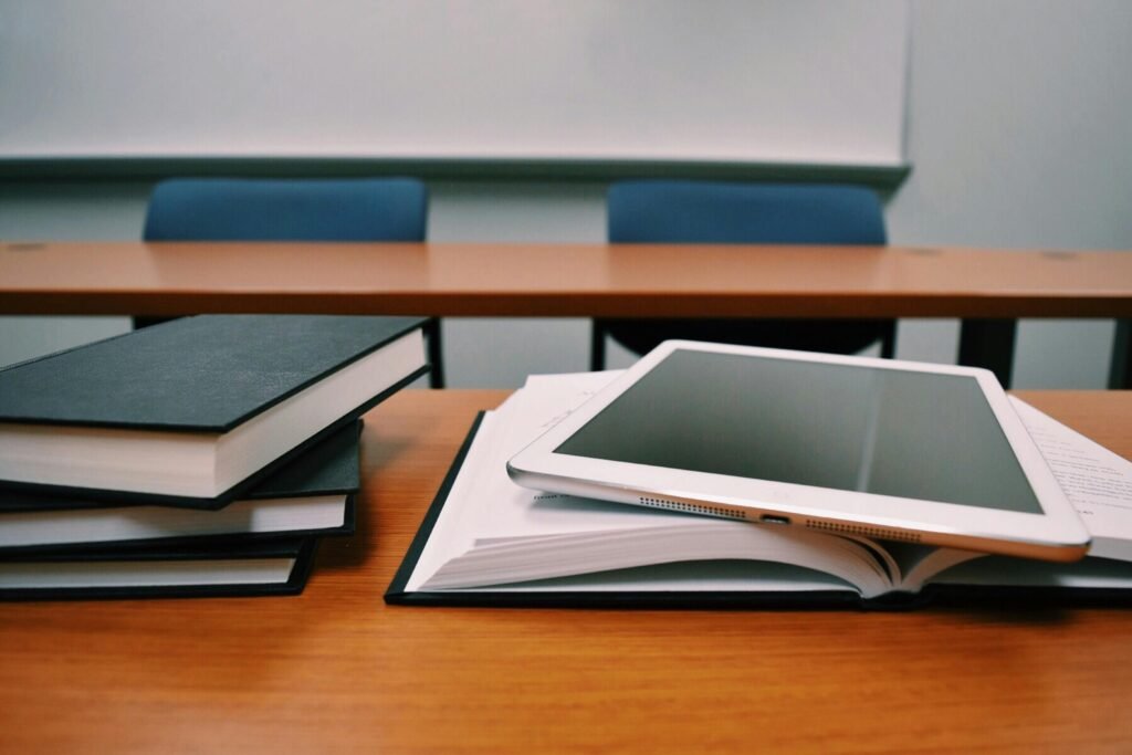 Stack of books and an open notebook with a tablet on a wooden classroom desk, with chairs and a whiteboard in the background.