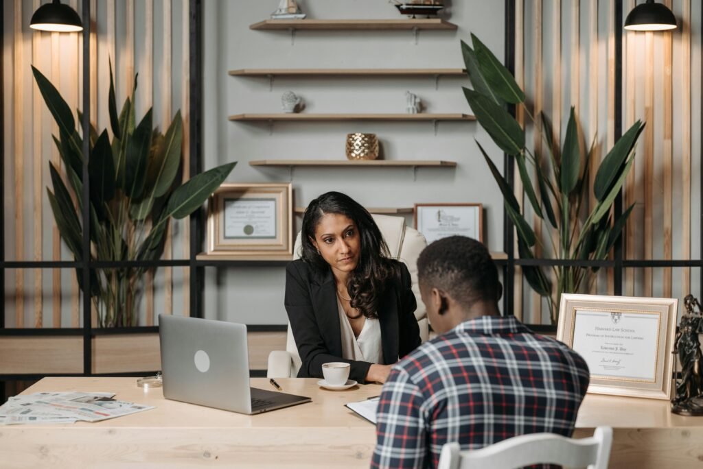 Two people sit across a desk in a modern office, engaged in a professional conversation with laptops and documents